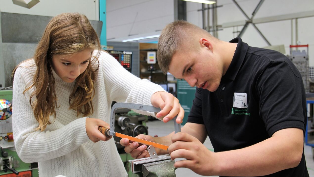 Auszubildender zeigt einer Praktikantin am Schraubstock das richtige Feilen während eines Praktikums